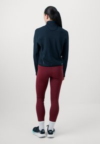 Mujer con coleta negra que lleva una chaqueta azul marino, leggins burdeos, calcetines blancos y zapatillas en blanco y negro, de pie mirando hacia atrás.