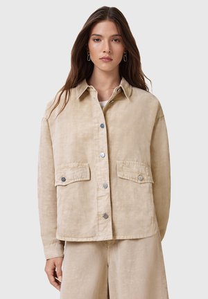 Beige jasje van textuurstof, met knopen aan de voorkant, twee borstzakken en een klassieke kraag, gedragen over een wit topje.