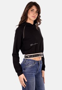 Hoodie nero cropped con cappuccio regolabile, cuciture accentate in oro e vita elastica con logo. Indossato con jeans blu.