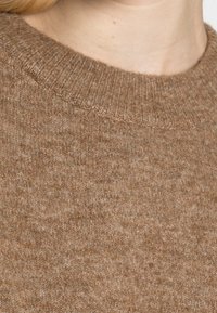 Nahaufnahme einer Person, die einen braunen gestrickten Pullover mit einem gerippten Rundhalsausschnitt trägt, wobei die helle Haut am Hals sichtbar ist.
