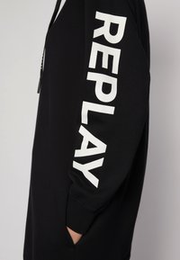 Svart långärmad plagg med vit fet "REPLAY" text tryckt vertikalt på ärmen och en hand som är stoppad i en sidficka.