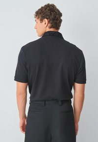 Camisa polo negra con cuello texturizado, manga corta y un corte relajado. Combinada con pantalones negros a medida con bolsillos traseros.