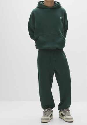 Dark green hoodie with a front pocket and drawstring hood, paired with matching relaxed-fit sweatpants. White logo detail on the chest.