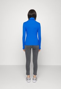 Jersey de manga larga, con cuello alto acanalado en azul brillante; diseño ceñido; combinado con vaqueros grises y zapatillas deportivas; visto desde atrás.