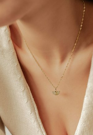 Goldene Halskette mit einem fächerförmigen Anhänger mit strukturierten Rillen, die an einer zarten Kette hängt, getragen über einem leichten, gestrickten Pullover.