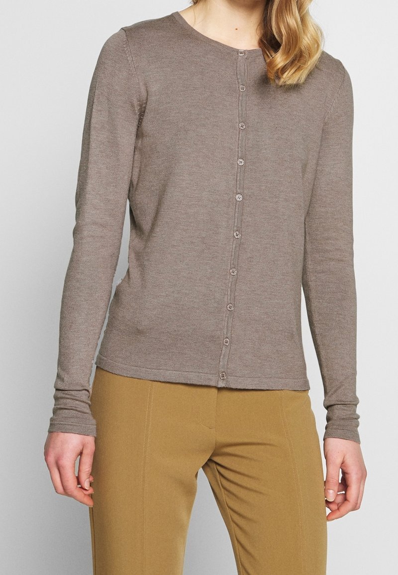 Femme portant un cardigan boutonné marron clair et un pantalon couleur moutarde, debout devant un fond uni.