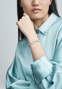 Srebrna zapestnica z zapletenimi okroglimi nastavki in prozornimi kamni. Nosi jo model z modro satenasto srajco.