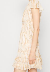 Femme portant une robe à manches courtes, beige et blanche avec un imprimé serpent et un ourlet rassemblé, se tenant devant un fond clair.