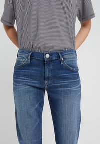 Siniset farkut, joissa on klassinen viisipaikkainen malli, edessä nappikiinnitys ja hieman kapeutuvat lahkeet. Kankaan tekstuuri ja haalistuneet yksityiskohdat.