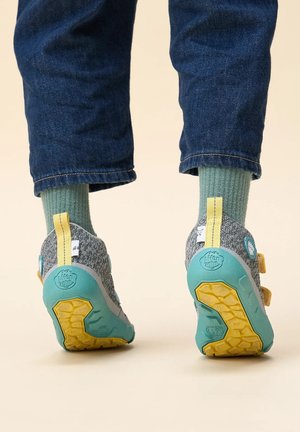 Szare i turkusowe buty gumowe z żółtymi akcentami i teksturowaną podeszwą, noszone z podwiniętymi niebieskimi dżinsami i zielonymi skarpetkami. Widoczne zapięcia na rzep.