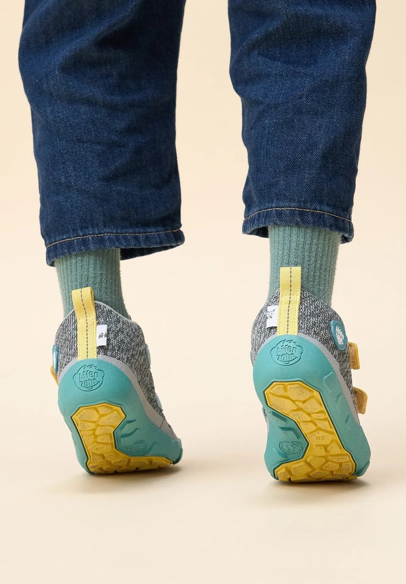 Scarpe di gomma grigie e teal con accenti gialli e suole testurizzate, indossate con jeans blu arrotolati e calze verdi. Straps in Velcro visibili.