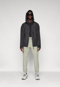 Veste imperméable noire avec capuche, associée à un pantalon cargo vert clair. Le modèle porte des lunettes de soleil réfléchissantes et des baskets multicolores.