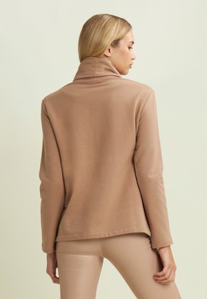 Maglione beige a maniche lunghe con colletto alto e orlo con spacco laterale, caratterizzato da una texture morbida e una vestibilità rilassata, indossato su leggings di colore chiaro.