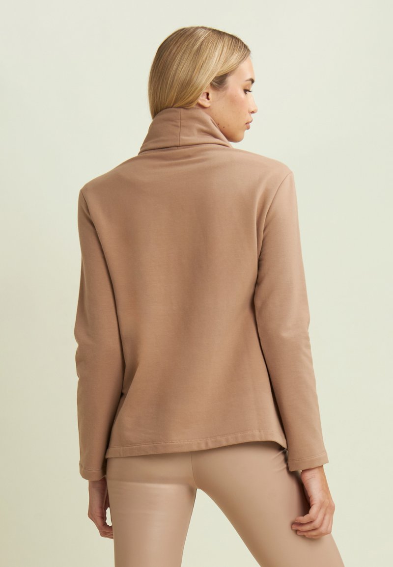 Maglione beige a maniche lunghe con colletto alto e orlo con spacco laterale, caratterizzato da una texture morbida e una vestibilità rilassata, indossato su leggings di colore chiaro.