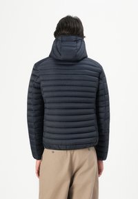 Man in een zwart gewatteerd hooded jack en beige broek, gezien van achteren tegen een eenvoudige witte achtergrond.