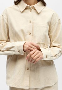 Chemise beige en velours côtelé avec fermeture à boutons sur le devant, col et poche poitrine unique. Tissu texturé et côtelé avec une coupe décontractée.