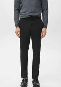 Mężczyzna ubrany w czarne wąskie spodnie, szarą koszulę z długim rękawem i czarne skórzane mokasyny, stoi na tle jasnego, gładkiego tła.