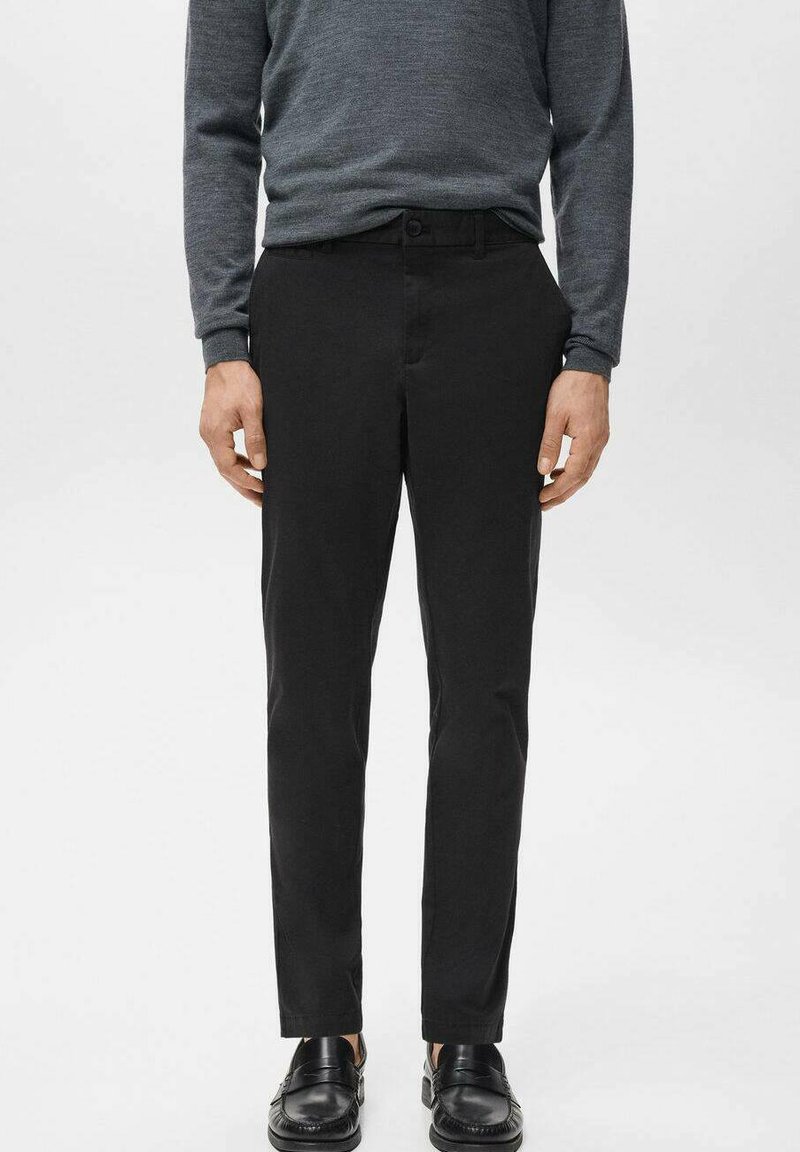 Mężczyzna ubrany w czarne wąskie spodnie, szarą koszulę z długim rękawem i czarne skórzane mokasyny, stoi na tle jasnego, gładkiego tła.