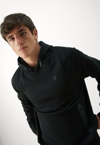 Schwarzer Hoodie aus weichem Material mit Kapuze und Kordelzug, ausgestattet mit einem kleinen Logo auf der Brust und mit Reißverschlusstaschen an den Seiten.