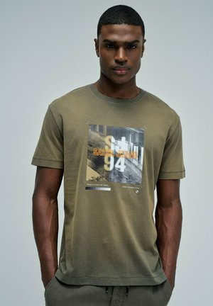 Joven con camiseta gráfica verde oliva con texto "SALSA JEANS 94" e imagen urbana, de pie con las manos en los bolsillos frente a un fondo liso.