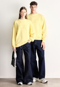 Κίτρινα βαμβακερά φούτερ με ανάγλυφο λογότυπο, σε συνδυασμό με σκούρα μπλε τζιν παντελόνια φαρδιάς γραμμής. Το μοντέλο κρατάει μια ασημί τσάντα. Casual στυλ.
