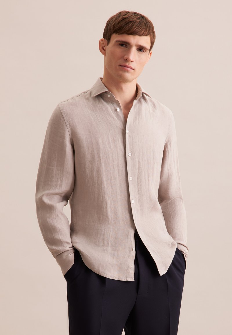 Camicia in lino beige chiaro con colletto button-down, tasca singola e maniche lunghe. Tessuto liscio e vestibilità rilassata. Abbinata a pantaloni scuri.