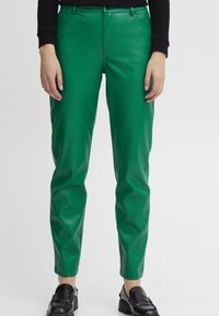 Persona con pantalones de cuero verde y mocasines negros, de pie con las manos relajadas a los lados sobre un fondo blanco liso.