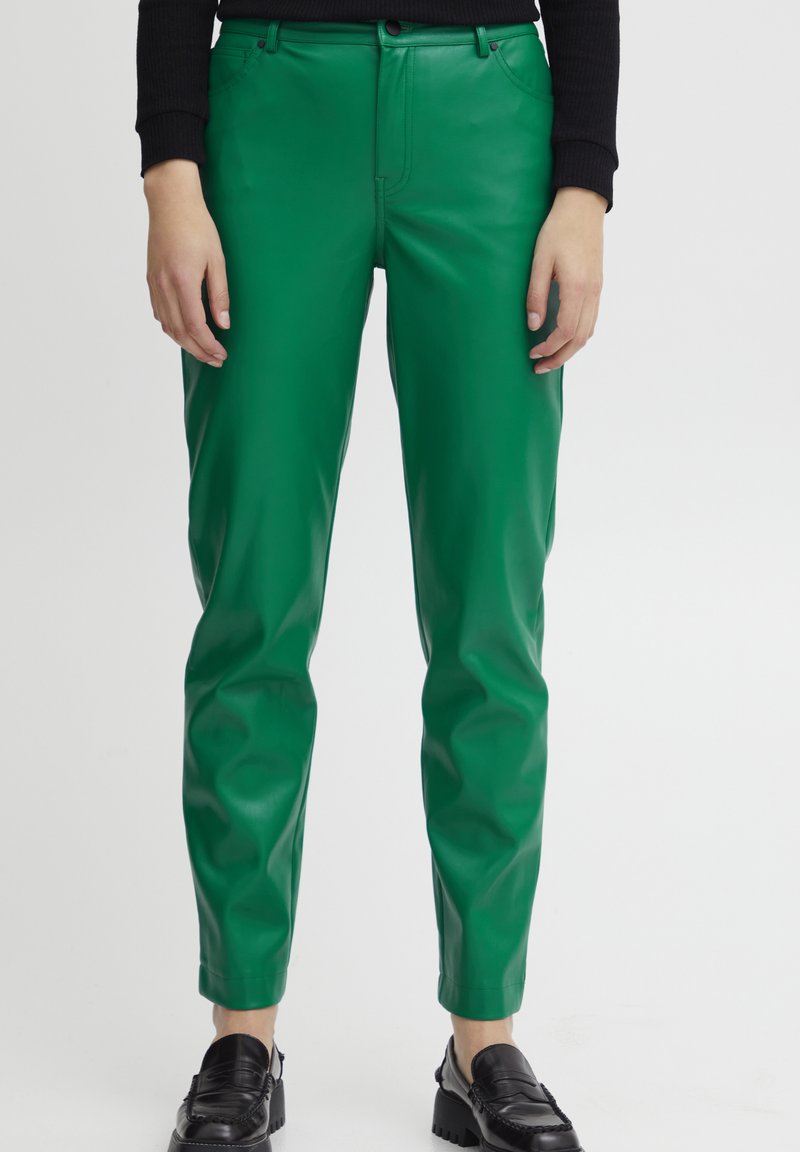 Persona con pantalones de cuero verde y mocasines negros, de pie con las manos relajadas a los lados sobre un fondo blanco liso.