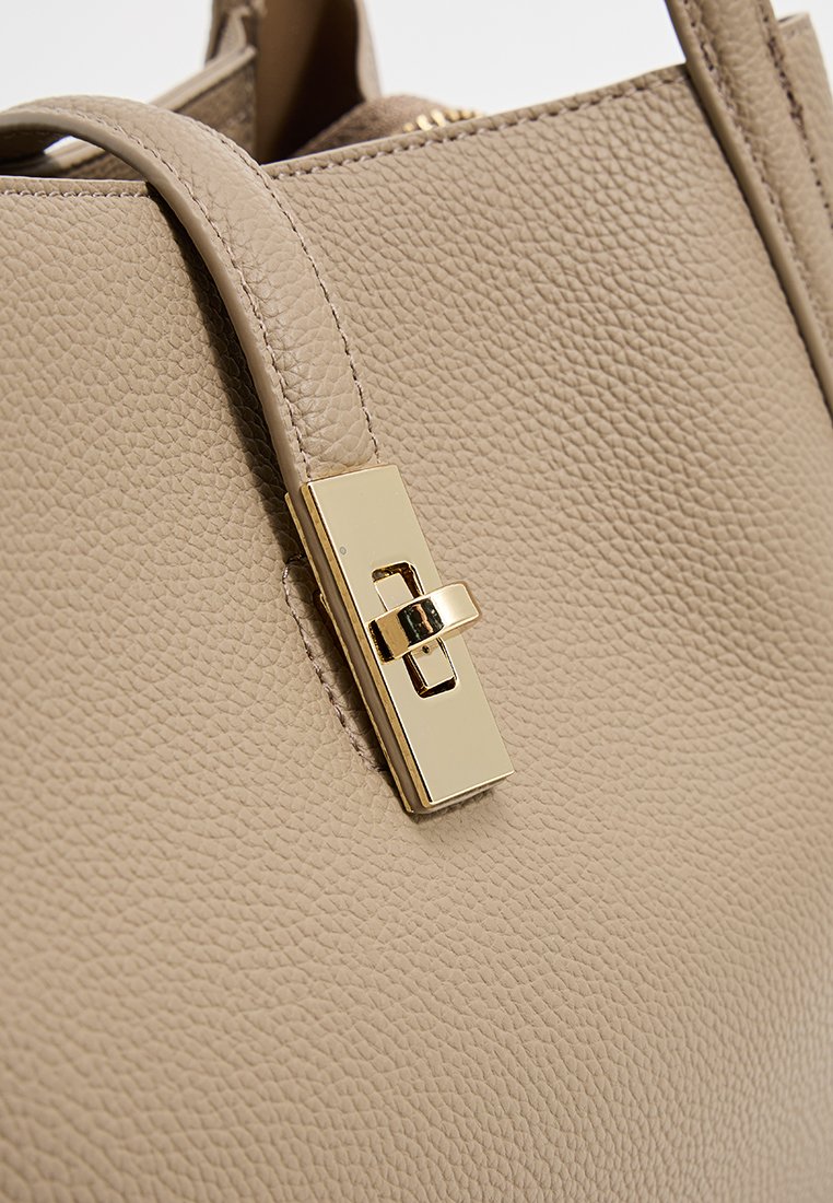 Beige leren handtas met een gestructureerd oppervlak, voorzien van een goudkleurige sluiting en stevige handvatten. Focus op het slanke rechthoekige ontwerp van de sluiting.