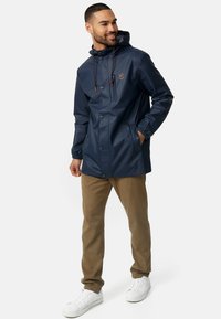 Marineblauwe waterdichte jas met capuchon, drukknoopsluiting, elastische manchetten en twee voorzakken; gecombineerd met kaki broek en witte sneakers.