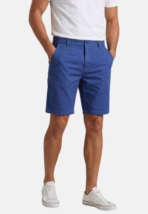 Homme portant un short bleu mi-genou, un t-shirt blanc et des baskets basses blanches, debout les mains dans les poches sur un fond blanc.