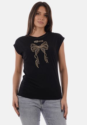 Donna che indossa un top nero senza maniche con design a fiocco dorato e testo "YES ZEE", abbinato a jeans grigio chiaro.