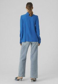 Niebieska bluzka z długim rękawem i wysokim kołnierzem, z teksturowanej tkaniny, z bufkami na ramionach, w połączeniu z jasnoniebieskimi szerokimi spodniami i butami na obcasie.