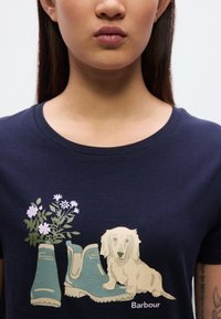 Μπλε ναυτικό μπλουζάκι με σχέδιο πράσινων λαστιχένιων μπότες, λευκά λουλούδια και εικονογράφηση ενός κρεμδαχλού ντα σκύλου. Περιλαμβάνεται το λογότυπο "Barbour".