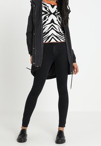 Personne portant une veste noire, un jean skinny noir, des chaussures noires et un pull à rayures zébrées noir et blanc avec une bordure orange.