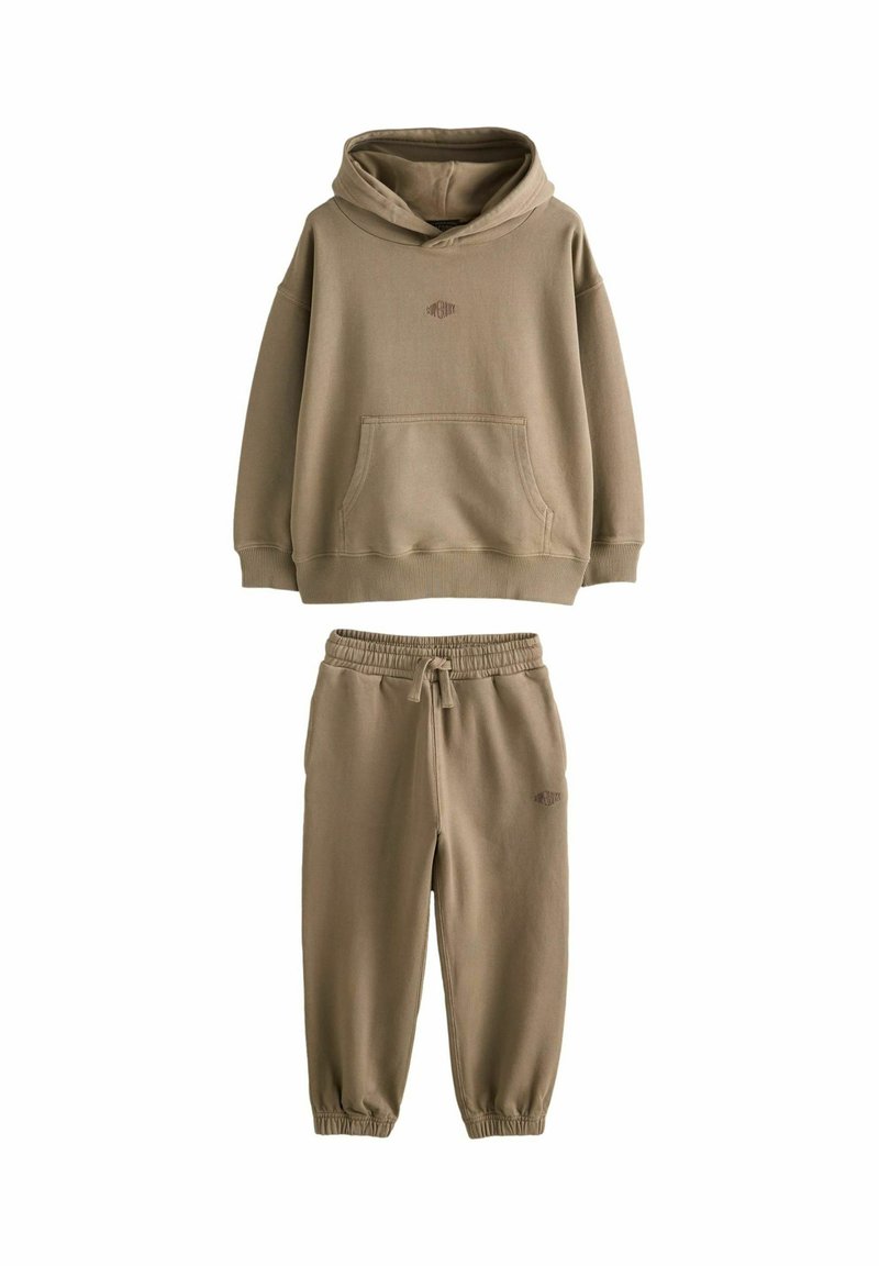 Hnedá mikina s kapucňou a zodpovedajúce jogger nohavice s elastickými manžetami a pásom so šnúrkou, s malým vyšitým logom na hrudi a stehne.
