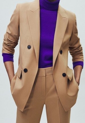 Kvinde iført beige dobbeltradet blazer og matchende bukser med lilla rullekravetrøje, hænderne i lommerne.