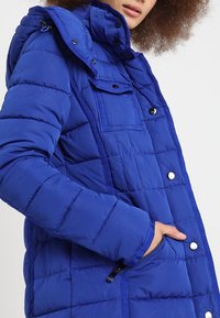 Mujer con una chaqueta acolchada azul con botones de presión y un bolsillo con cremallera, con la mano dentro del bolsillo, mostrando los detalles de la chaqueta.