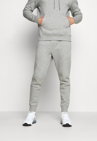 Grau meliertes Baumwollmix-Hoodie mit Kängurutasche, kombiniert mit passenden, schmal geschnittenen Jogginghosen. Weiße Sportschuhe mit schwarzen Akzenten.