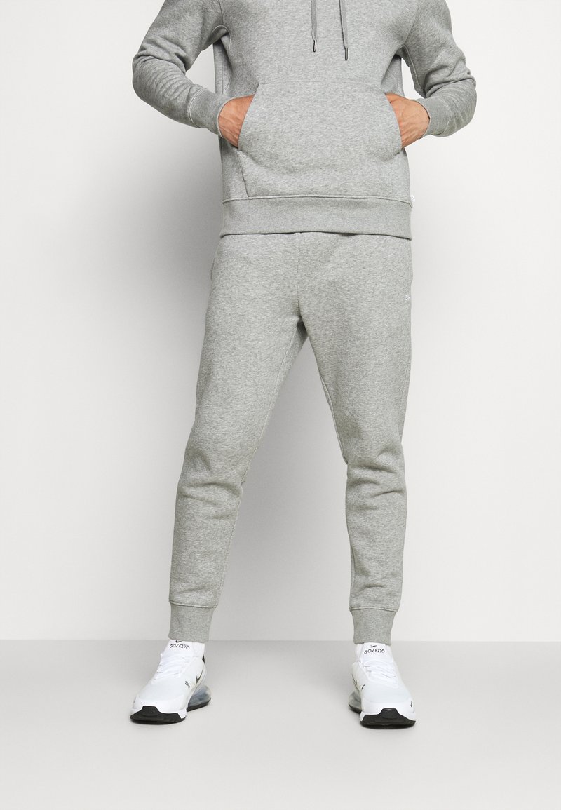Grau meliertes Baumwollmix-Hoodie mit Kängurutasche, kombiniert mit passenden, schmal geschnittenen Jogginghosen. Weiße Sportschuhe mit schwarzen Akzenten.
