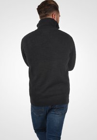 Dunkelgrauer, gestrickter Pullover mit hohem Kragen und geripptem Saum. Verfügt über strukturierte Details an den Ärmeln. Getragen mit blauen Jeans.