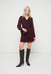 Bordowa sukienka kopertowa z długimi, przezroczystymi rękawami i wiązaniem w pasie, zestawiona z czarnymi kozakami za kolano o ostrym czubku oraz małą czarną torebką na ramię.