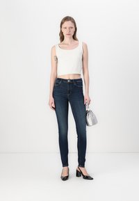 Skinny dark denim jeans with golden stitching, paired with a white cropped tank top and black heeled shoes. Accessorized with a silver handbag.