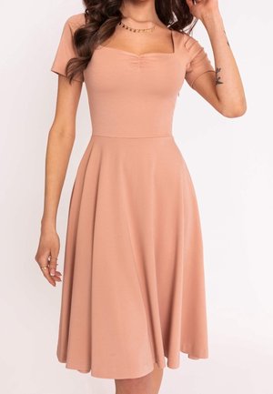 Femme portant une robe midi pêche à manches courtes, avec un décolleté carré froncé et une jupe évasée, montrant un tatouage sur son avant-bras.