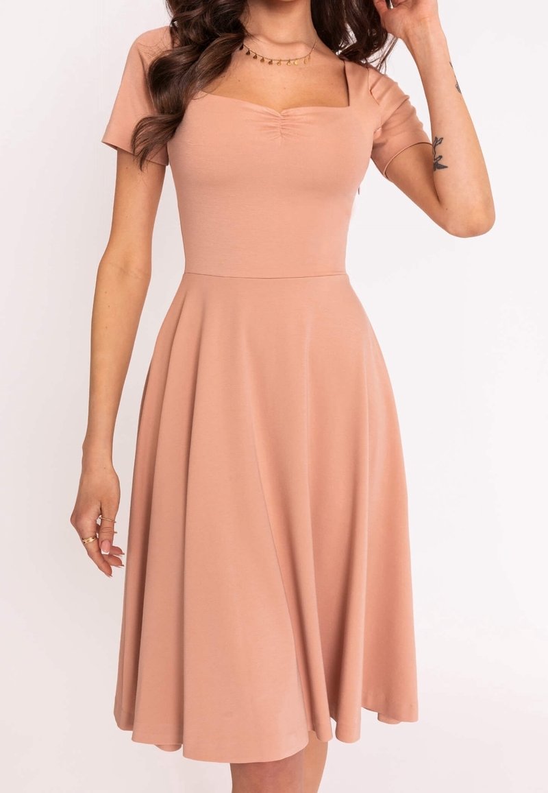 Femme portant une robe midi pêche à manches courtes, avec un décolleté carré froncé et une jupe évasée, montrant un tatouage sur son avant-bras.