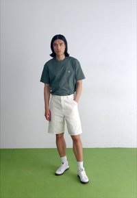 T-shirt vert à manches courtes, shorts blancs et baskets blanches. Le modèle se tient sur un sol vert avec un arrière-plan minimaliste. Design simple, coupe décontractée.