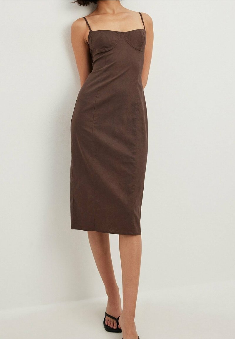 Femme portant une robe midi marron cintrée, sans manches, avec fines bretelles et des talons ouverts noirs sur un fond blanc uni.