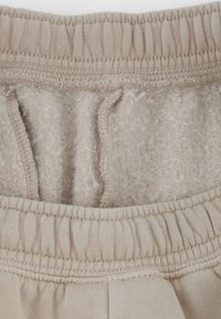 Fascia beige dei pantaloni della tuta con elastico e cordino. L'interno è foderato con un tessuto morbido e peloso. Dettagli delle cuciture visibili lungo i bordi.