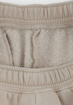 Beige tailleband van joggingbroek met elastiek en trekkoord. De binnenkant is gevoerd met zachte, pluizige stof. Naaddetails zichtbaar langs de randen.