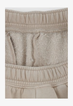 Fascia beige dei pantaloni della tuta con elastico e cordino. L'interno è foderato con un tessuto morbido e peloso. Dettagli delle cuciture visibili lungo i bordi.
